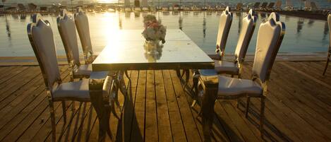 A wooden deck with a table and chairs, overlooking a pool and the ocean.