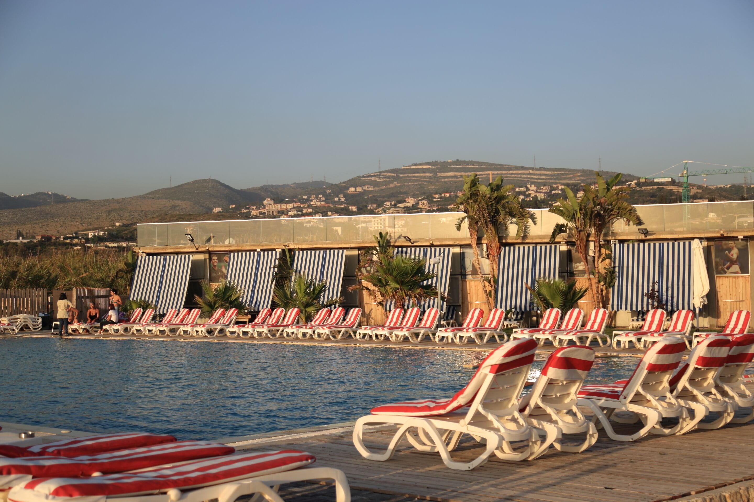 Un área junto a la piscina con sillones de descanso y sombrillas, cielo despejado y una vista distante de colinas.
