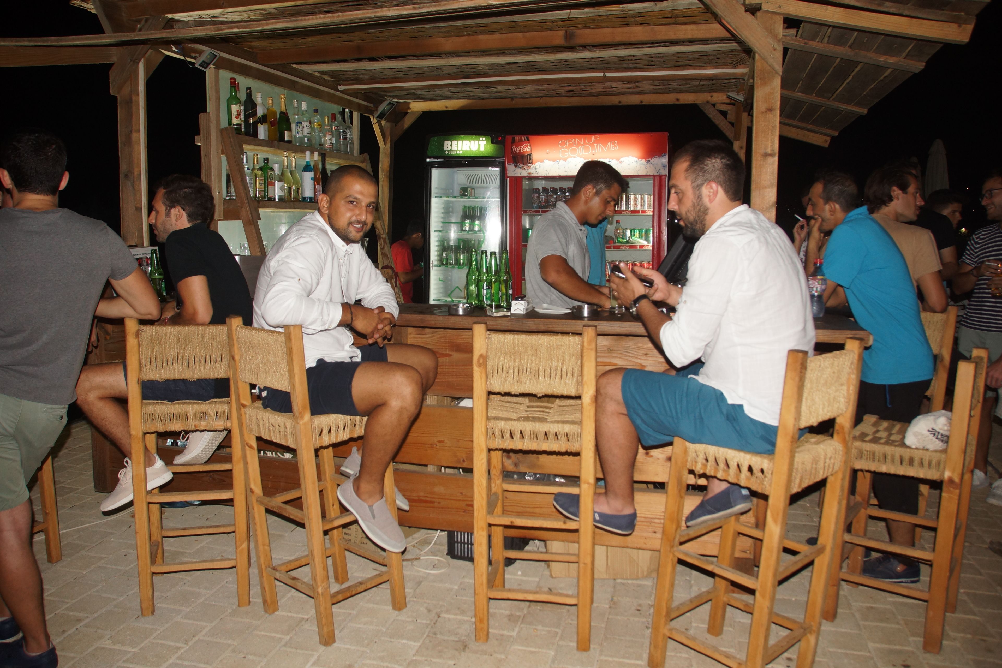 Zona de bar con sillas y mesas de madera, un refrigerador lleno de bebidas y una barra con una persona sirviendo tragos.