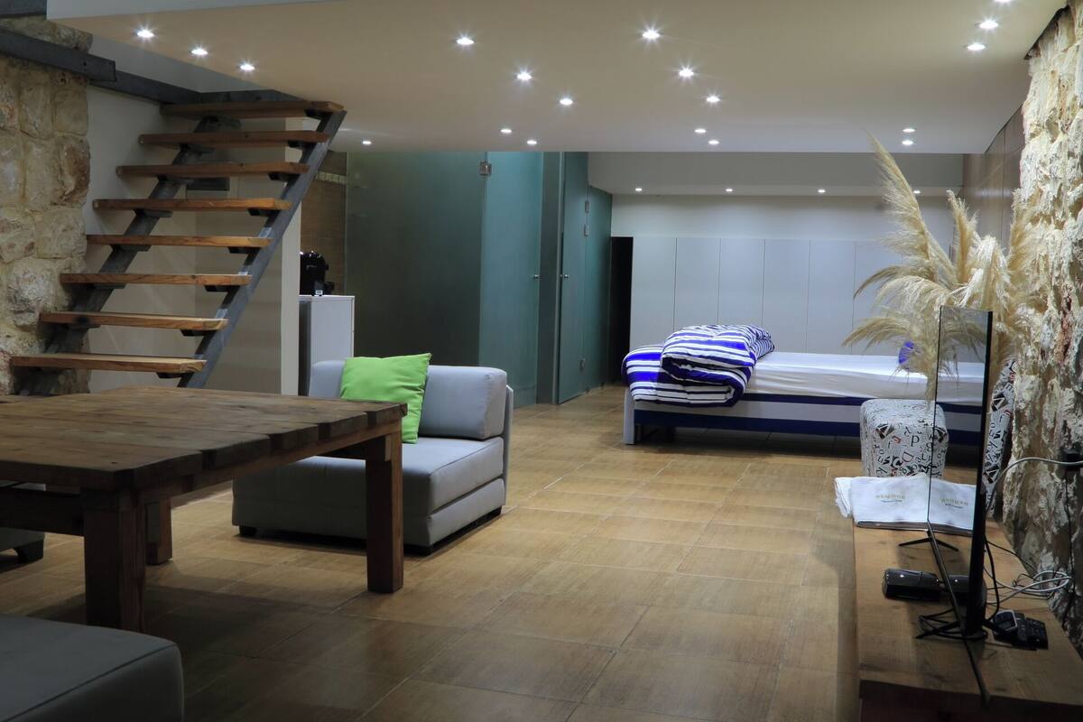 Un vaste espace de vie comprenant une table en bois, un canapé, un lit avec un coussin à rayures et un escalier.