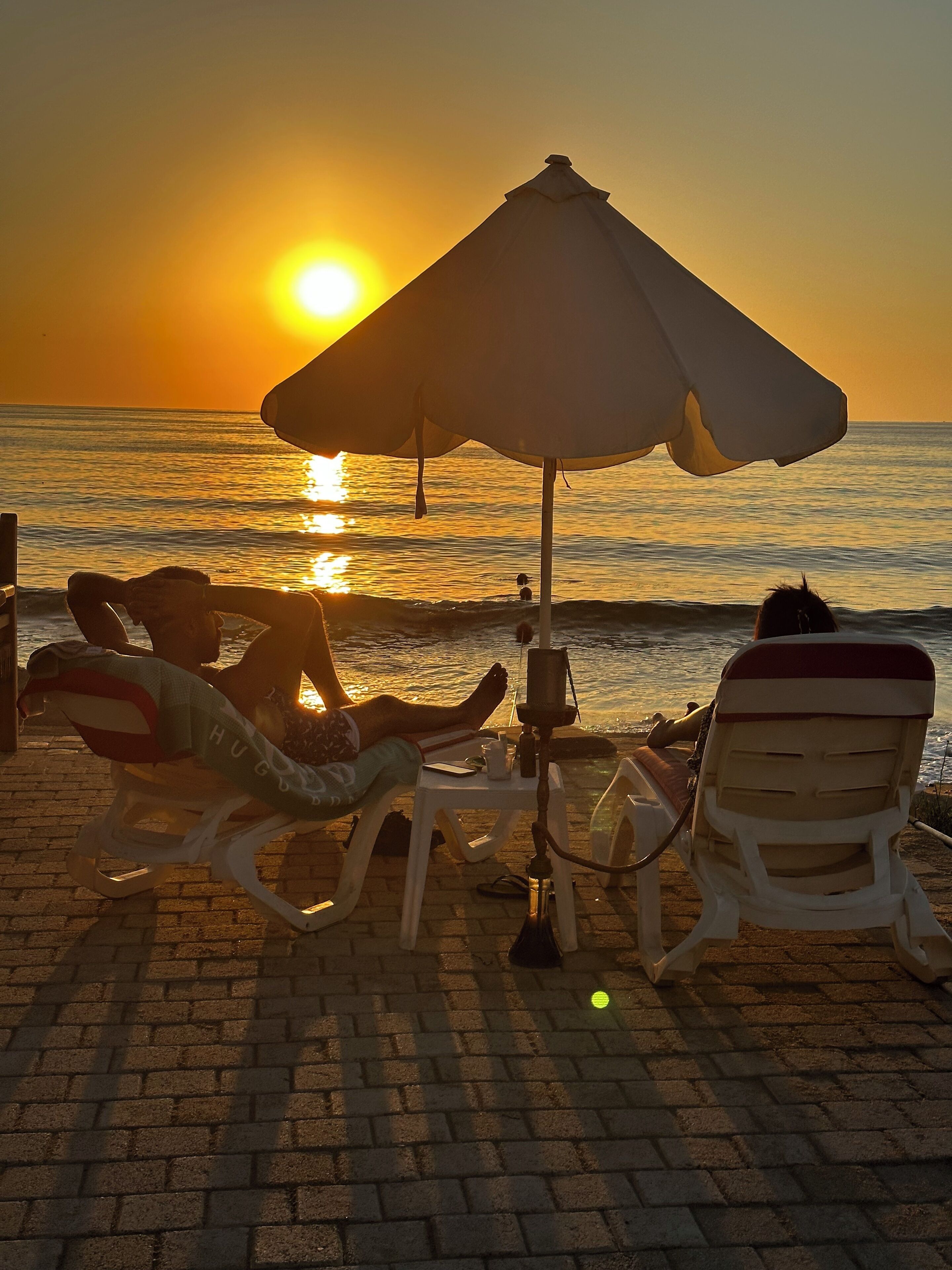 A beach scene with two lounge chairs under a large umbrella, a person relaxing, and the sun setting over the ocean.