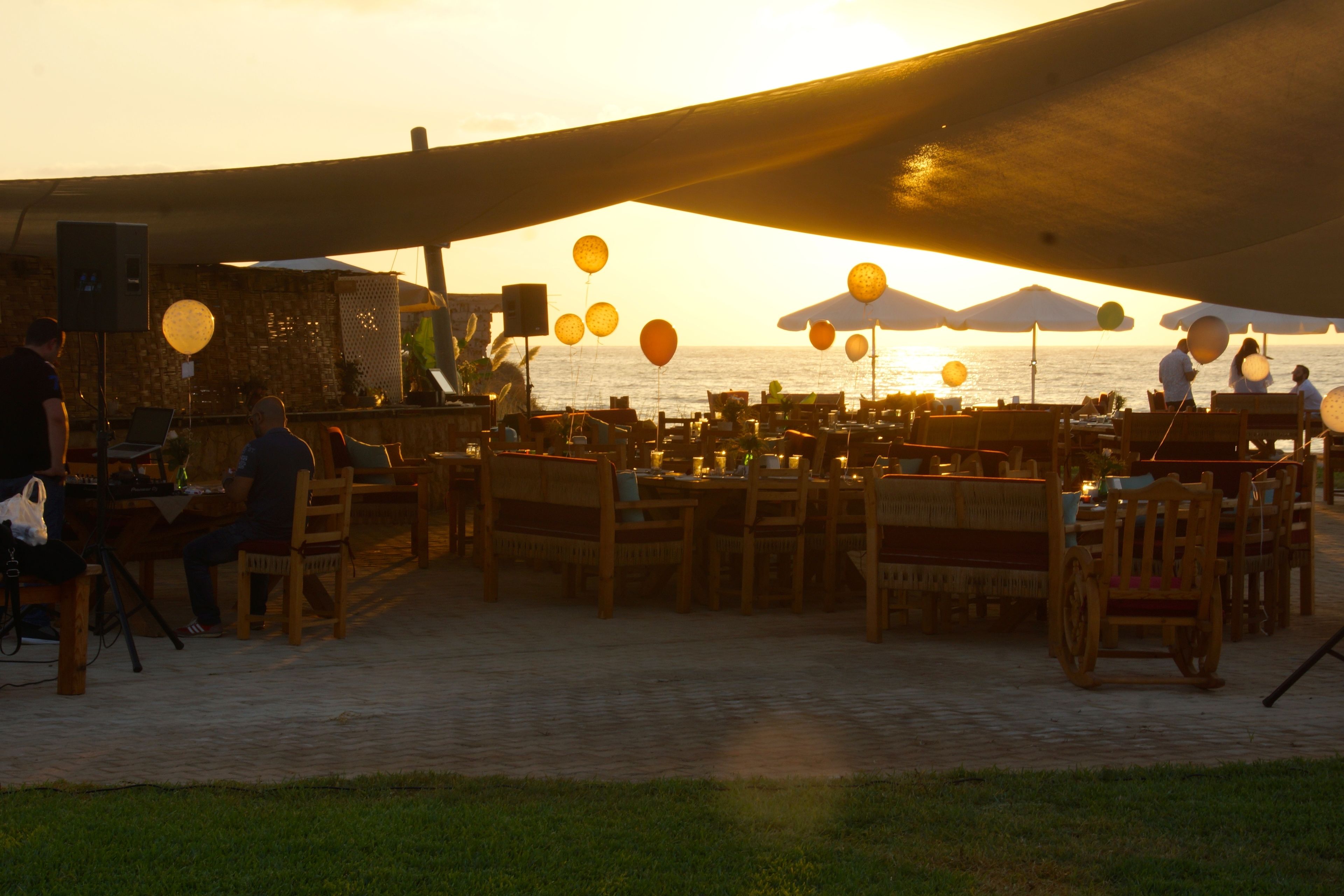An outdoor dining area with tables and chairs, a speaker, and a view of the sea at sunset.