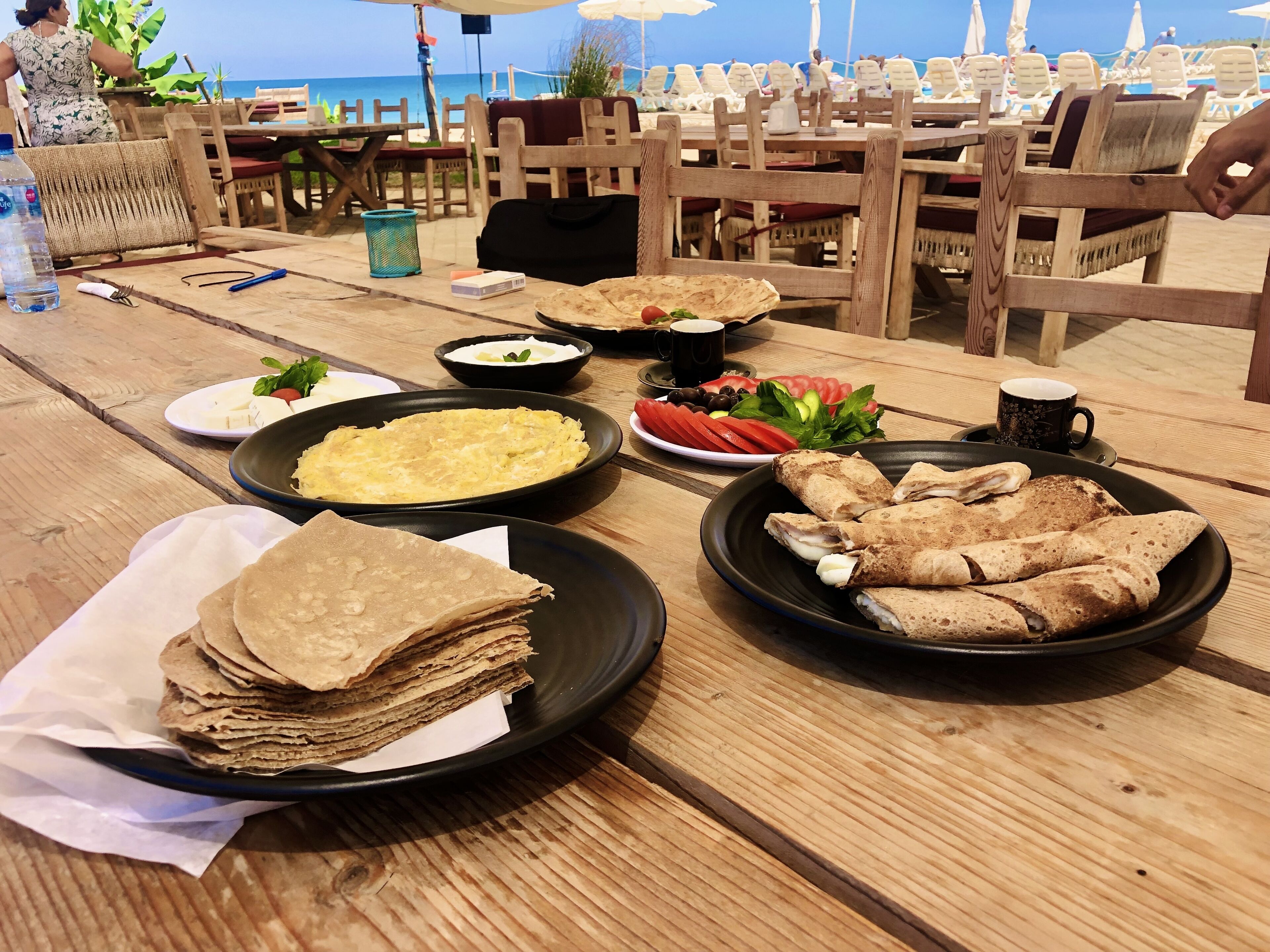 A wooden table with various food items, including a stack of crepes, a plate of sliced tomatoes, and a bowl of soup.