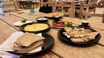 A wooden table with various food items, including a stack of crepes, a plate of sliced tomatoes, and a bowl of soup.