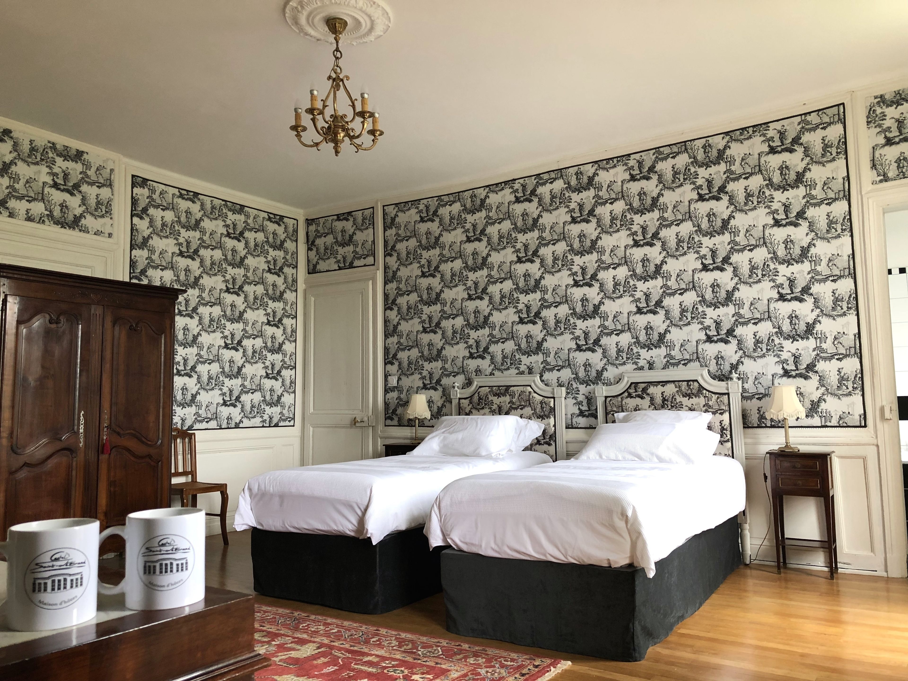 A bedroom with two beds, a chandelier, and patterned wallpaper.