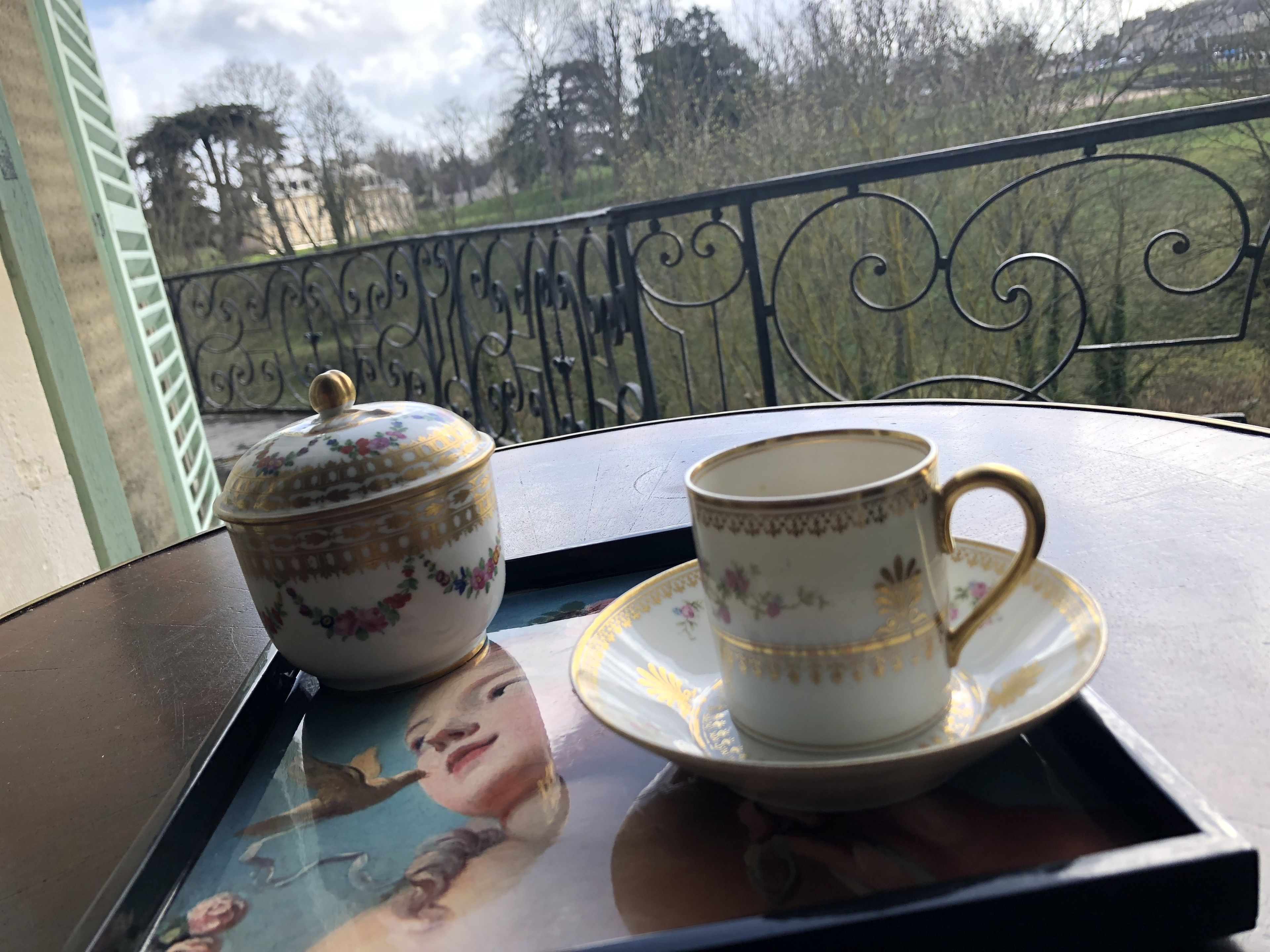 A table with a teapot and cup on a tray, with a view of trees and buildings in the background.
