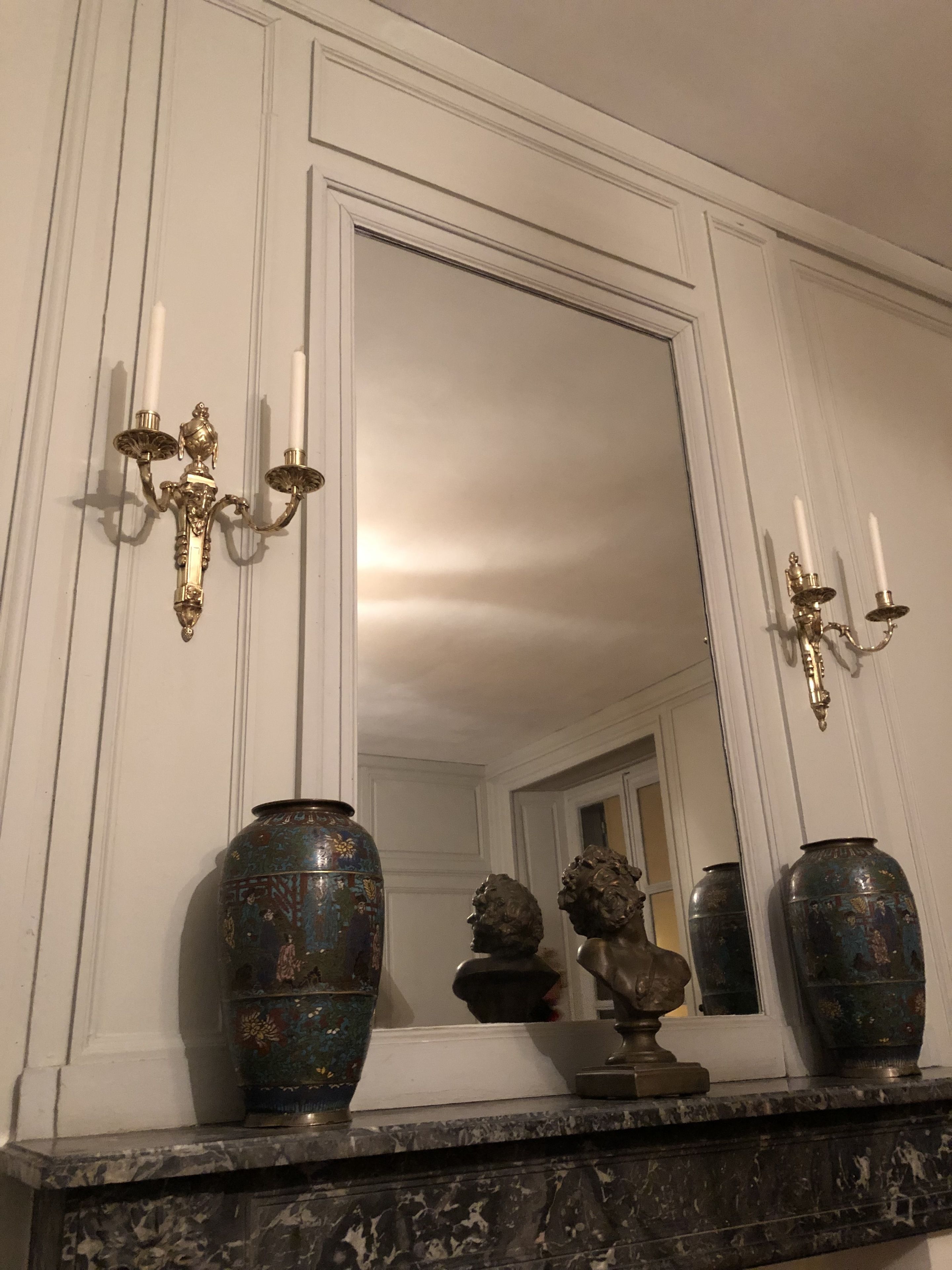 A room with a large mirror, two ornate wall sconces, and two decorative vases on a marble mantelpiece.