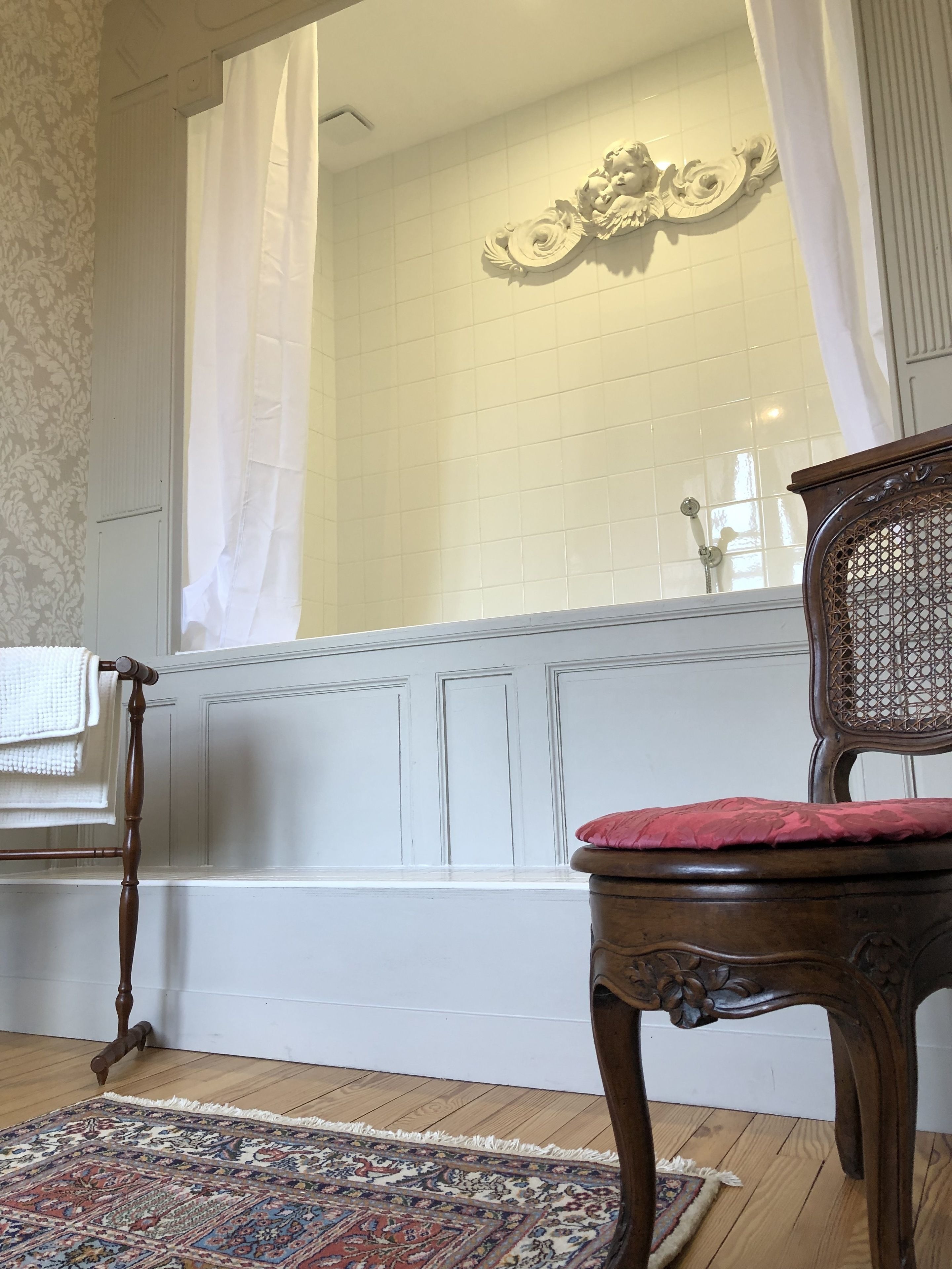 Een badkamer met een badkuip, een houten stoel met een rode kussentje, een tapijt met een patroon en een decoratief wandobject.