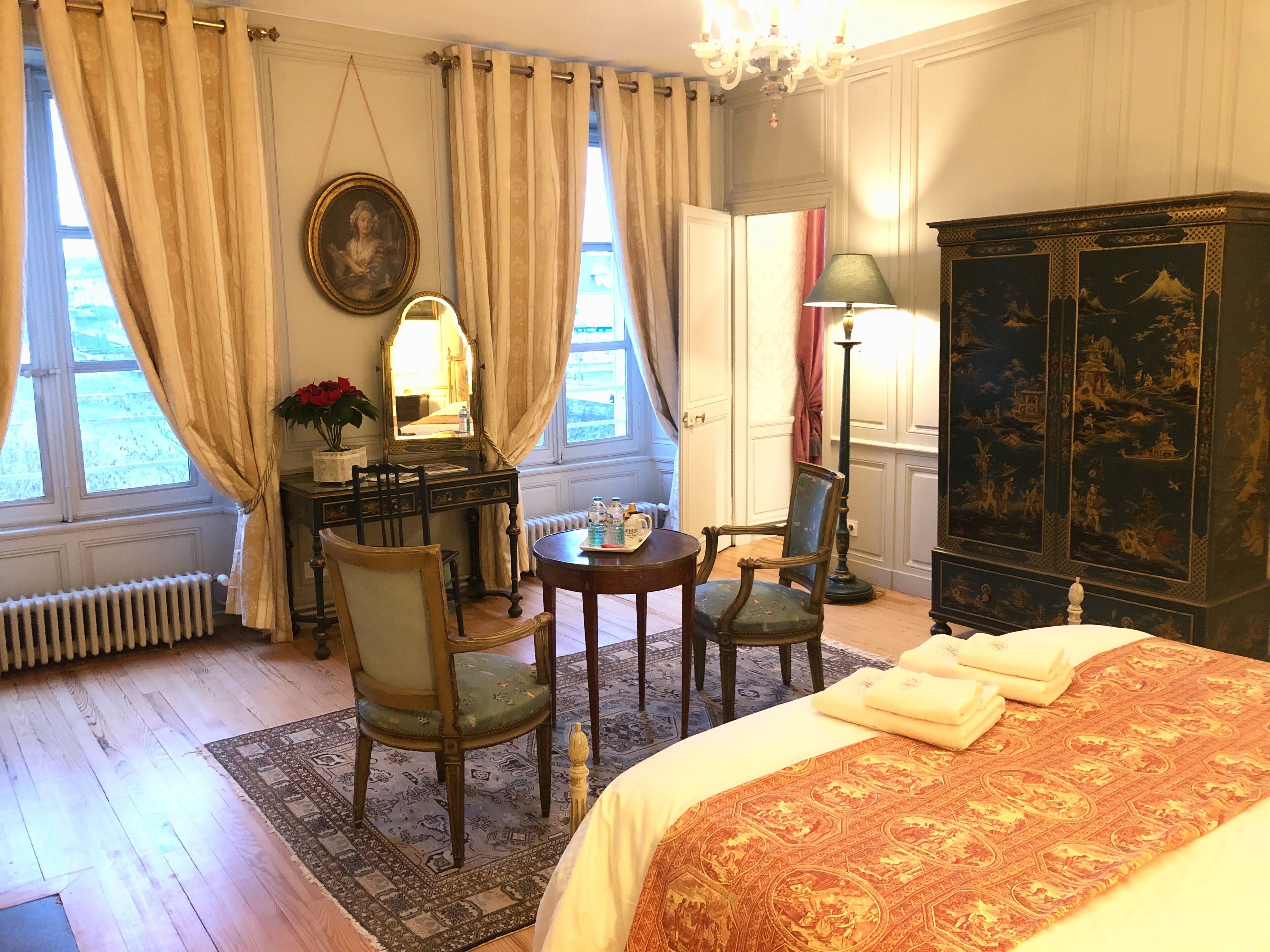 A hotel room with a bed, two chairs, a desk with a lamp, a dresser, and a large window with curtains.