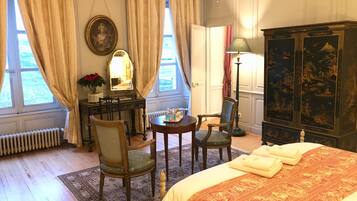 A hotel room with a bed, two chairs, a desk with a lamp, a dresser, and a large window with curtains.