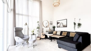 Una sala de estar moderna con un sillón gris, un sofá negro, una mesa de centro y una guitarra. Hay plantas, un espejo y cuadros en las paredes.