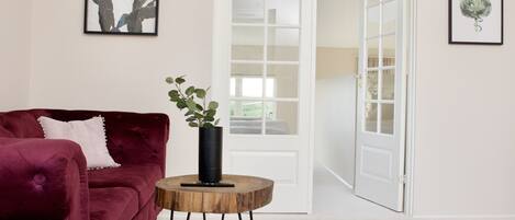 A room with a red velvet sofa, a small wooden side table with a plant, and a white door leading to another room.