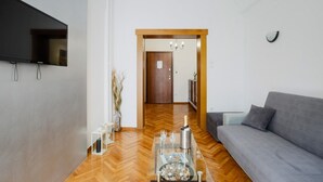 A modern living room with a grey sofa, a flat-screen TV mounted on the wall, a wooden door leading to another room, and a glass coffee table with a bottle and glasses.