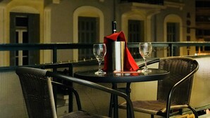 A table with two chairs, a bottle, and two wine glasses on a balcony with a railing.