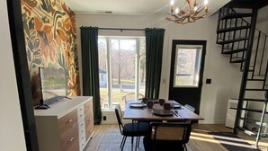 Uma sala de jantar com mesa, cadeiras e um candelabro. Escada à direita e porta que dá para o exterior.