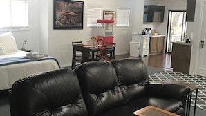 Un salon avec un canapé en cuir noir, une petite table en bois et un coin repas avec une table et des chaises rouges. Il y a également un coin cuisine avec des placards et une fenêtre.