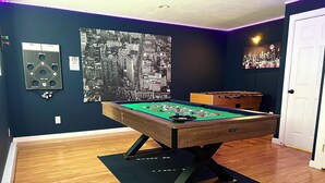 Una mesa de billar en una habitación con paredes oscuras, un cartel con un paisaje urbano y suelo de madera.