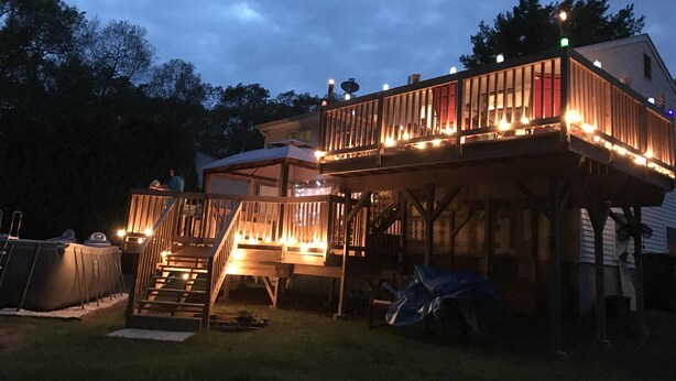 Una terraza de dos pisos con escalera y barandillas de madera, rodeada por un césped y una tienda de campaña.