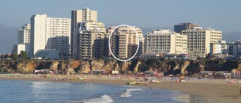 A coastal cityscape with high-rise buildings, a sandy beach, and a rocky cliff.
