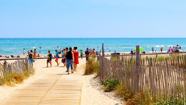 Ein hölzerner Steg führt zu einem Strand, auf dem Menschen spazieren und sonnenbaden.