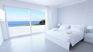 A modern bedroom with a large bed, white bedding, a nightstand, and a view of the ocean.