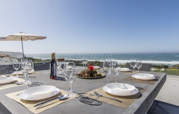 Mesa de comedor al aire libre con platos, vasos y cubiertos, con vista al océano.