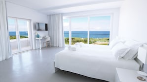 A modern hotel room with a large bed, a desk, and a TV. The room has large windows with a view of the sea and greenery.