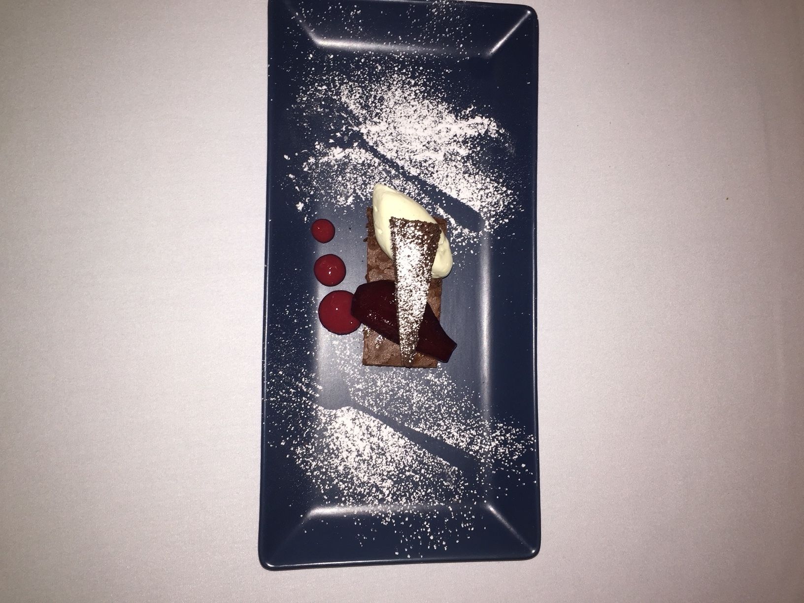 A dessert on a rectangular plate with powdered sugar, chocolate, and red spheres.