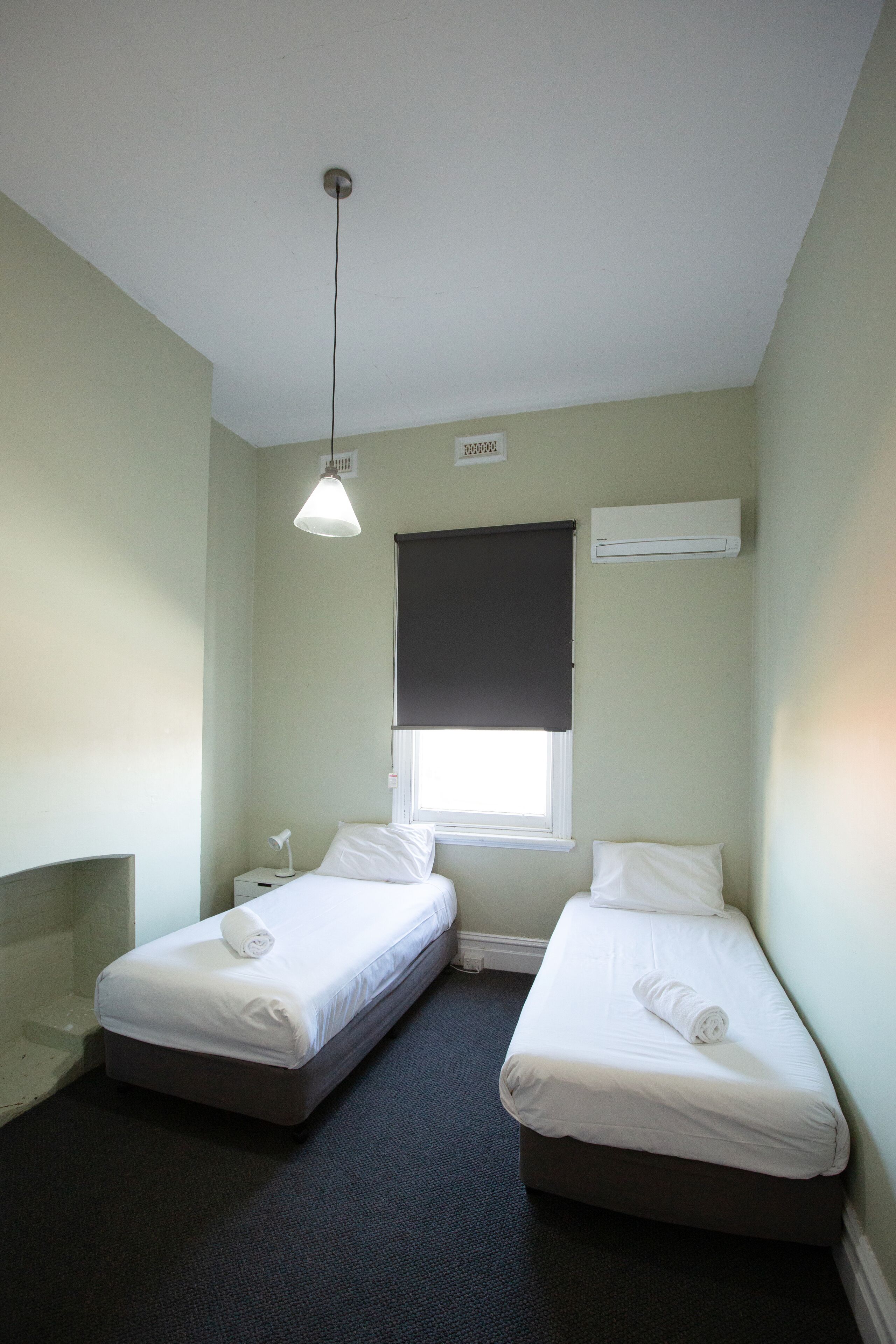 Una habitación pequeña y limpia con dos camas individuales, una ventana con cortina, una lámpara en el techo y un equipo de aire acondicionado.