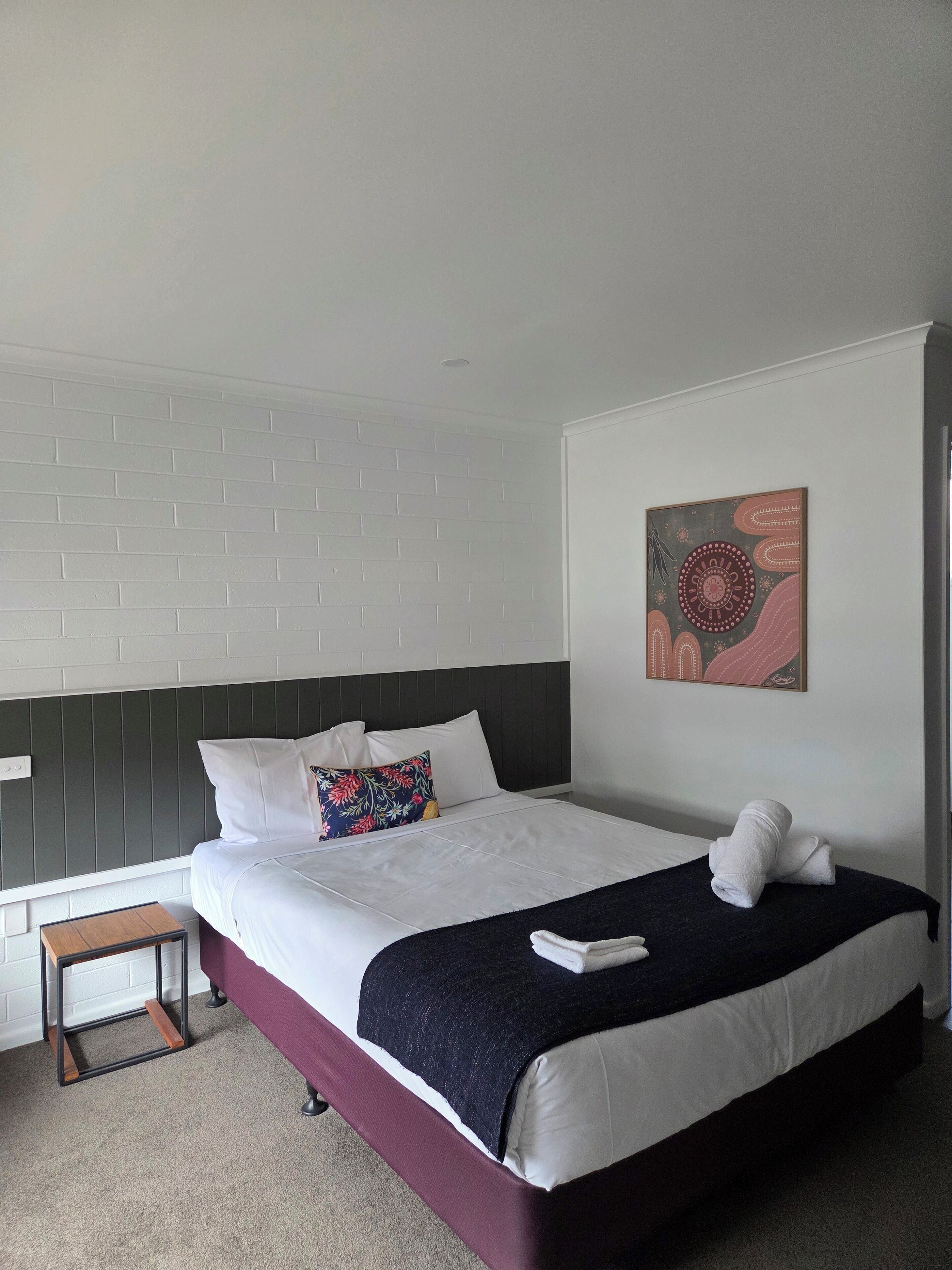 Quarto de hotel bem organizado, com uma cama, uma pequena mesa de cabeceira de madeira, uma obra de arte pendurada na parede e uma parede de tijolos brancos.