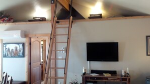 Een kamer met een houten ladder, een flatscreen televisie, een bureau met een computer en een deur naar een andere kamer.