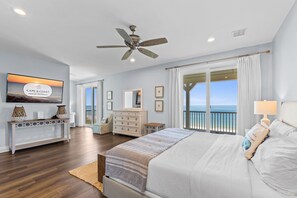 A bedroom with a bed, a dresser, a vanity, a ceiling fan, and a large window with a view of the ocean.
