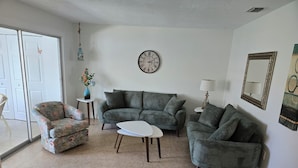 A living room with a sofa, armchair, and coffee table. There is a clock on the wall and a vase with flowers on a side table.