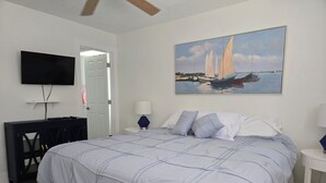A bedroom with a large bed, a television on the wall, a painting of sailboats, and a desk with a lamp.