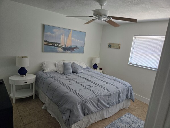 A bedroom with a bed, two bedside tables, a ceiling fan, a painting, a window with blinds, and a small rug.
