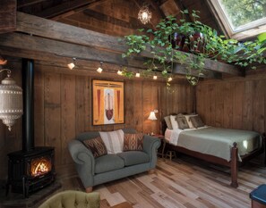 A cozy room with a sofa, a bed, a wooden floor, a wall-mounted painting, a hanging plant, a lantern, and a fireplace.