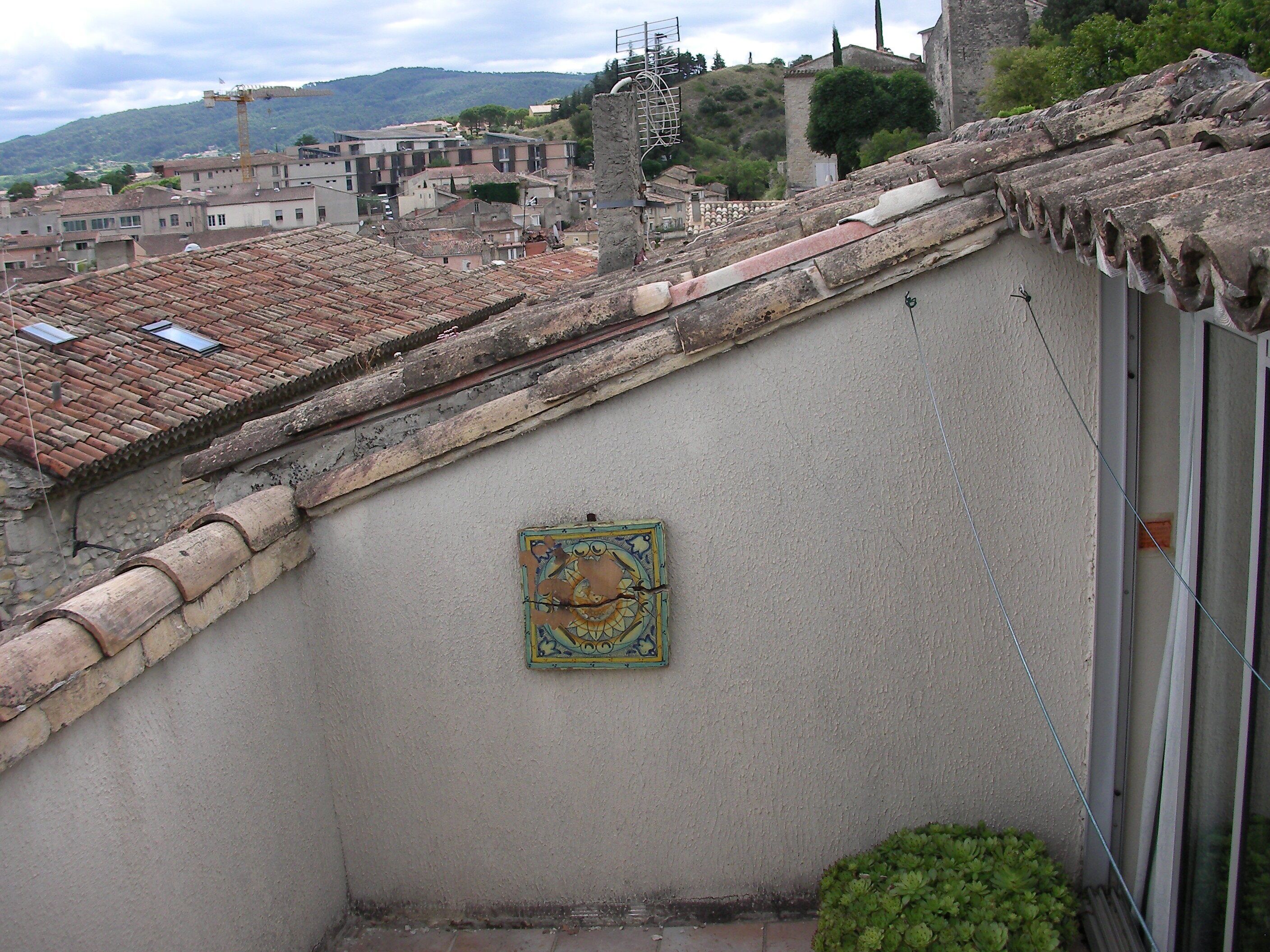 Un toit-terrasse recouvert de tuiles, orné d’une petite mosaïque décorative, et offrant une vue sur une ville avec ses bâtiments et les collines en arrière-plan.
