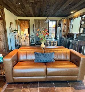 A brown leather sofa with a blue pillow, a vase with flowers, and a rustic kitchen with wooden cabinets and a dining area.