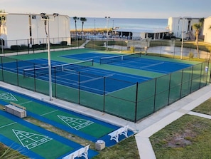 Un campo da tennis con superficie verde e blu, circondato da una recinzione, adiacente a una zona piscina.