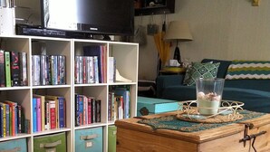 A living room with a television, bookshelves, a wooden chest, and a patterned rug. - Cozy 2 Bedroom Cottage Walking Distance to the Beach (Zurich)