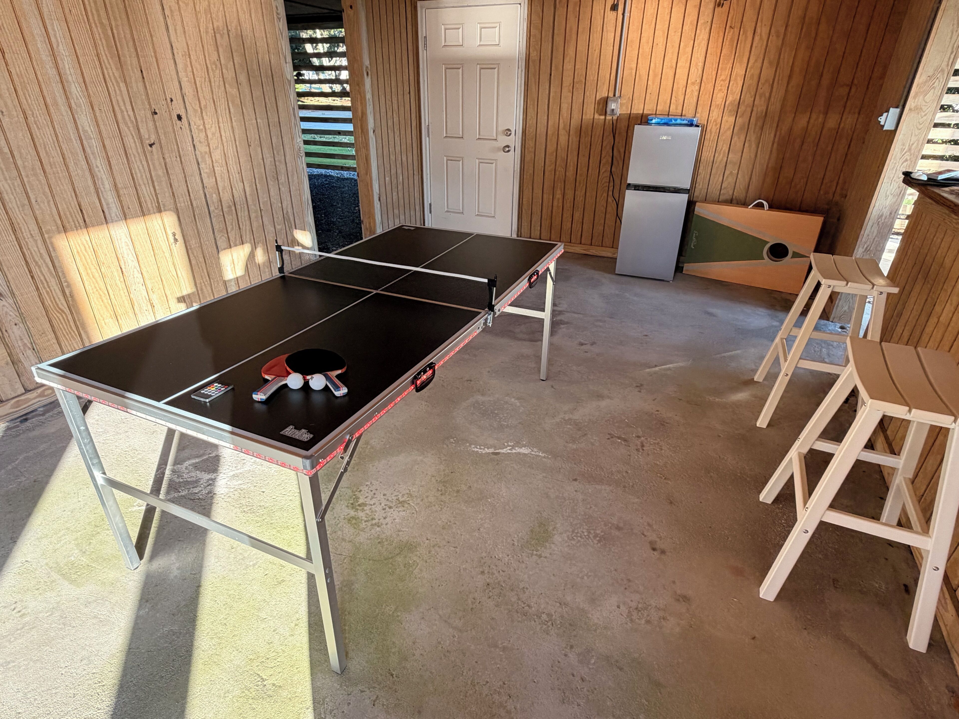A ping pong table with two chairs, a refrigerator, and a door in a wooden room.