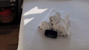 Una cama con una colcha blanca acolchada, dos almohadas, una toalla enrollada y un neceser.