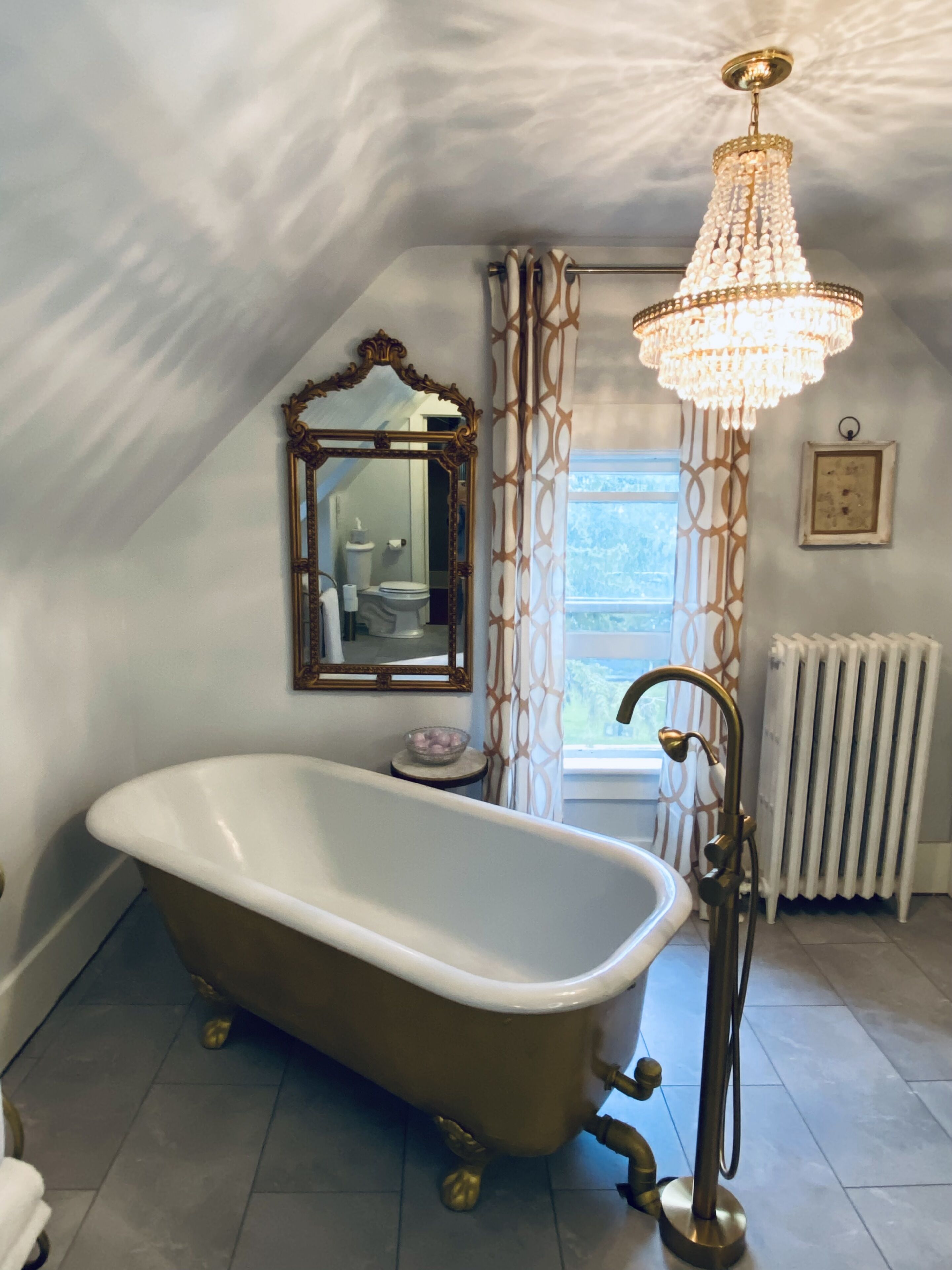 Een badkamer met een vrijstaand bad, een kroonluchter, een spiegel en een raam met gordijnen.