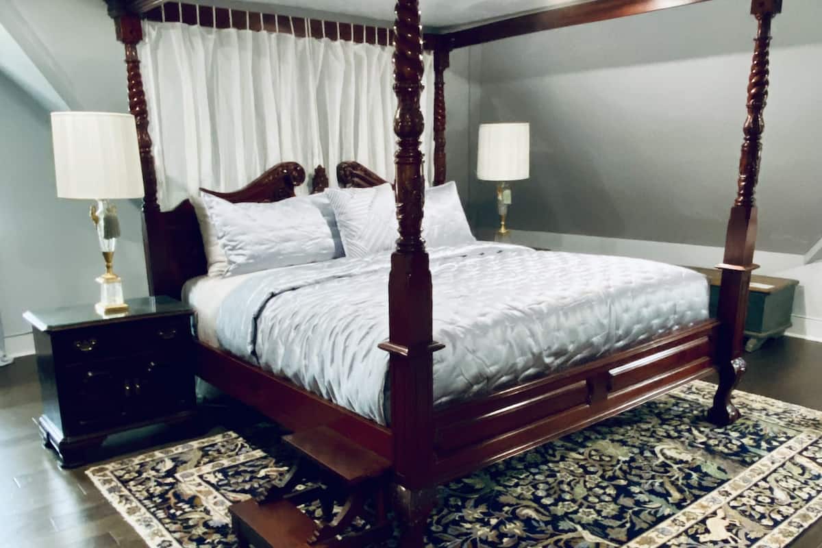 A four-poster canopy bed with a dark wooden frame and white bedding, positioned on a patterned rug in a room with a neutral color scheme.