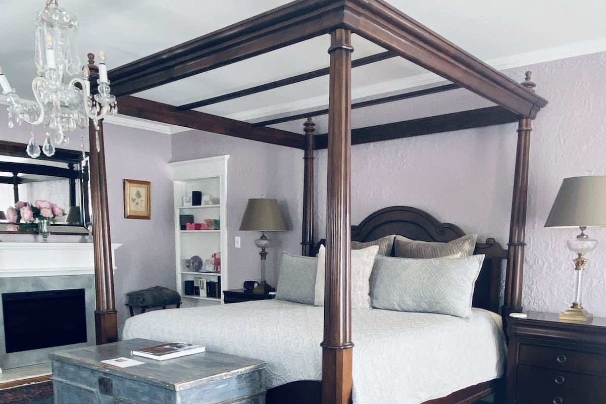 A four-poster bed with a wooden frame, a bedside table, a nightstand, a dresser, a mirror, a chandelier, and a patterned carpet.