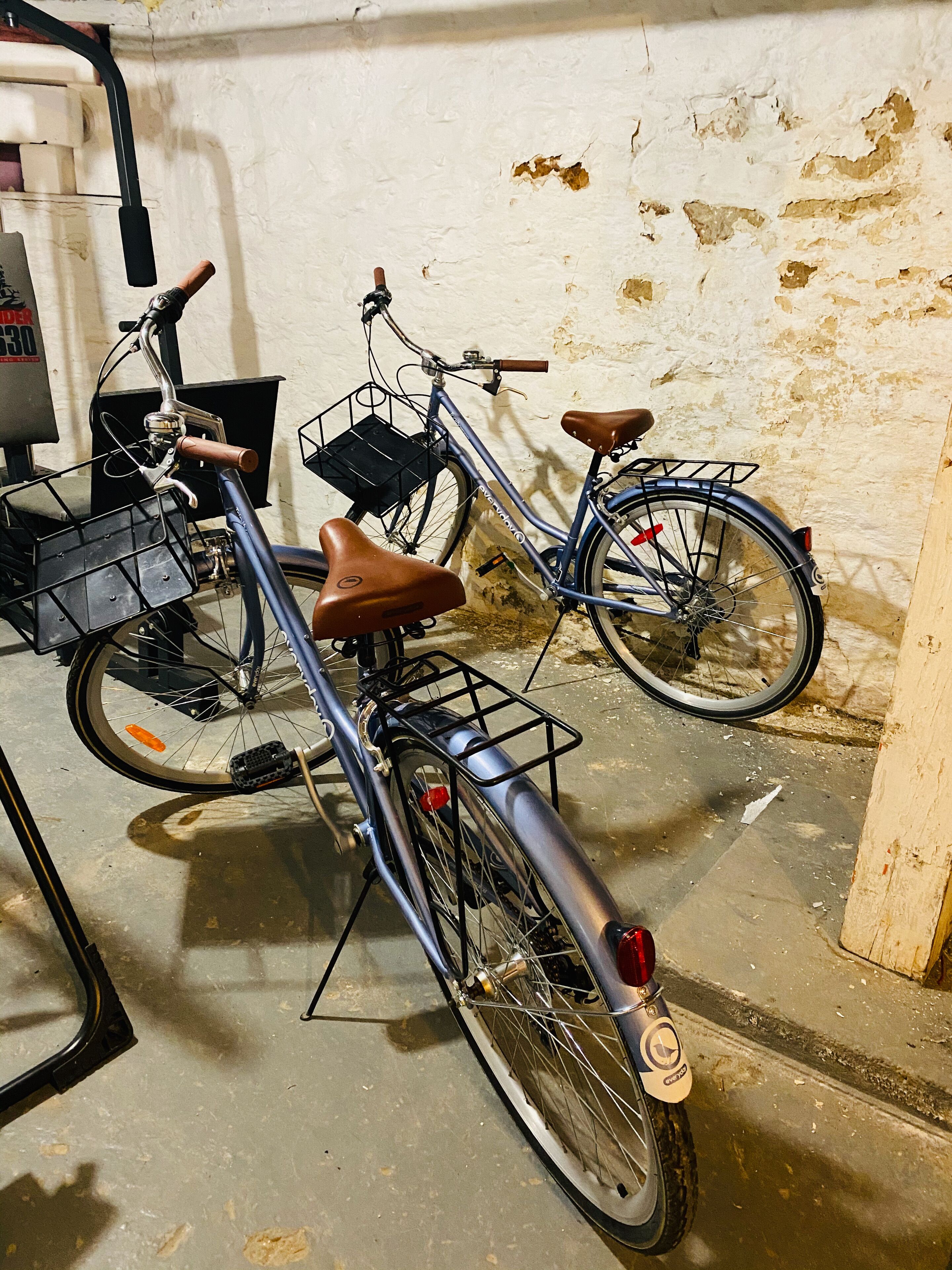 Ein Fahrrad mit Korb und Sitz, abgestellt in einem Innenraum mit Betonboden und einer weißen Wand mit abblätternder Farbe.
