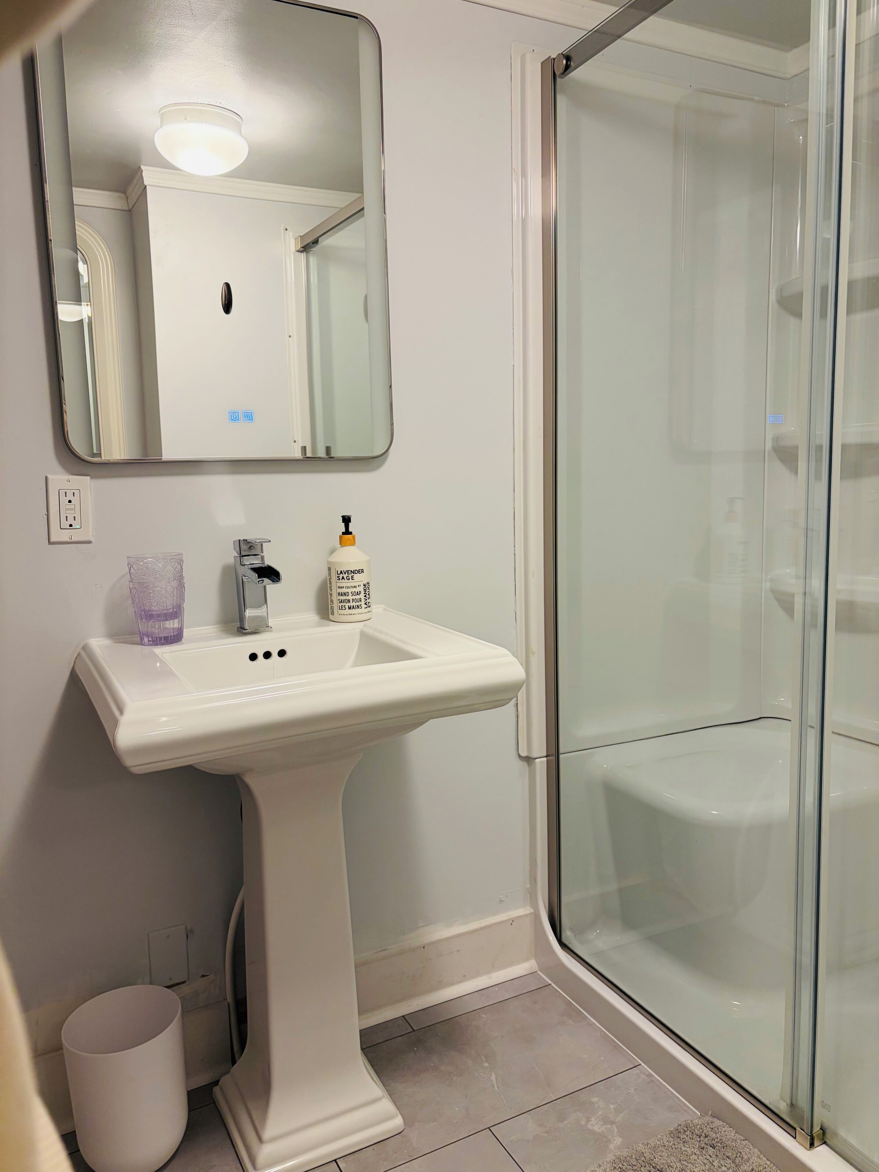 A bathroom with a white sink, a mirror, a glass shower enclosure, and a trash bin.