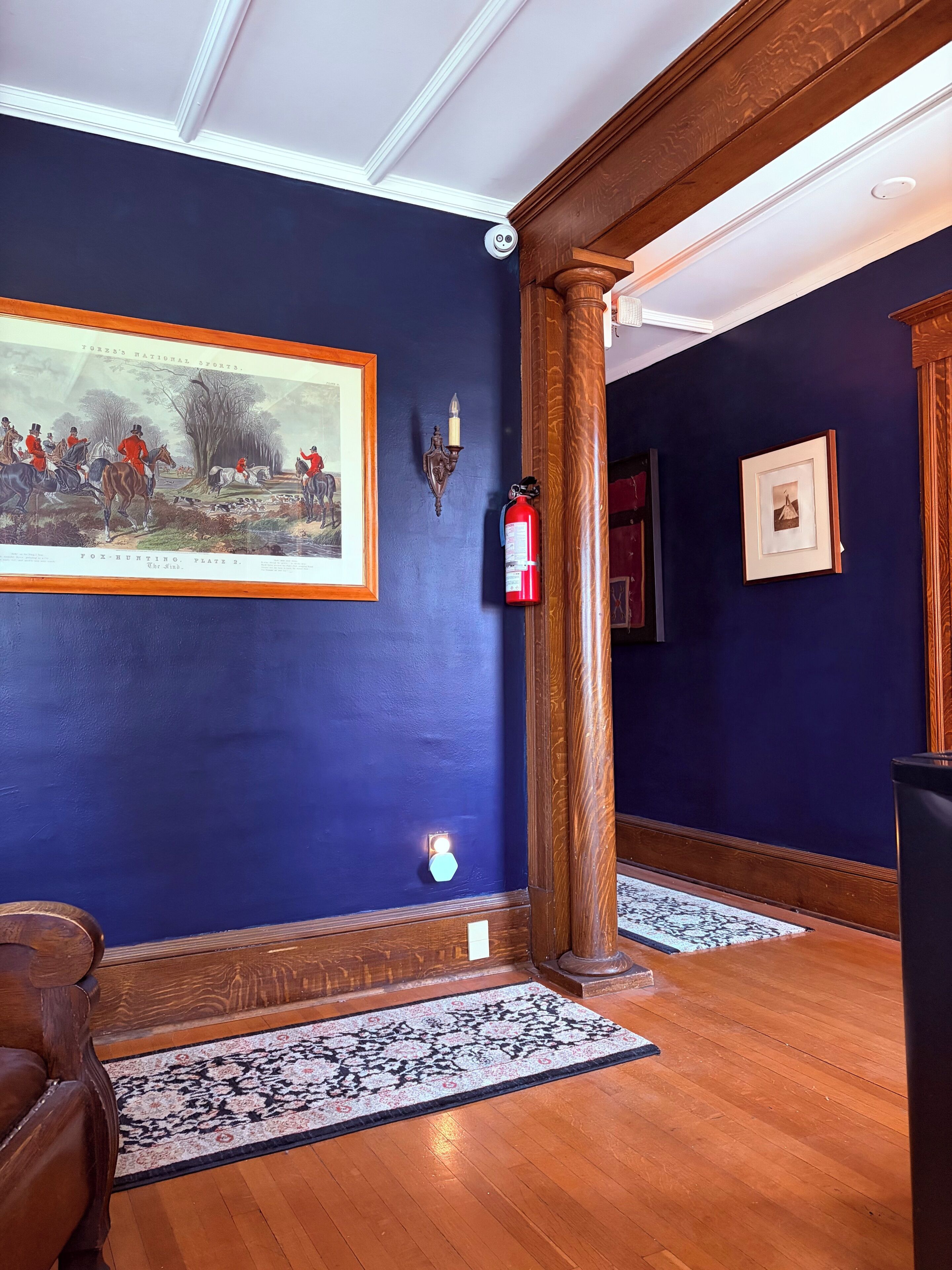 A hallway with dark blue walls, a framed historical sports illustration, a fire extinguisher, and a patterned rug.