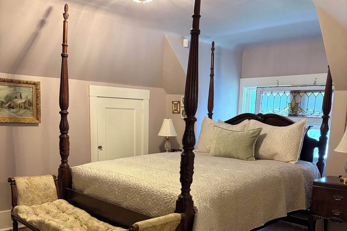 A four-poster bed with a canopy, a chaise lounge, a wooden nightstand, and a large window with a stained glass design.