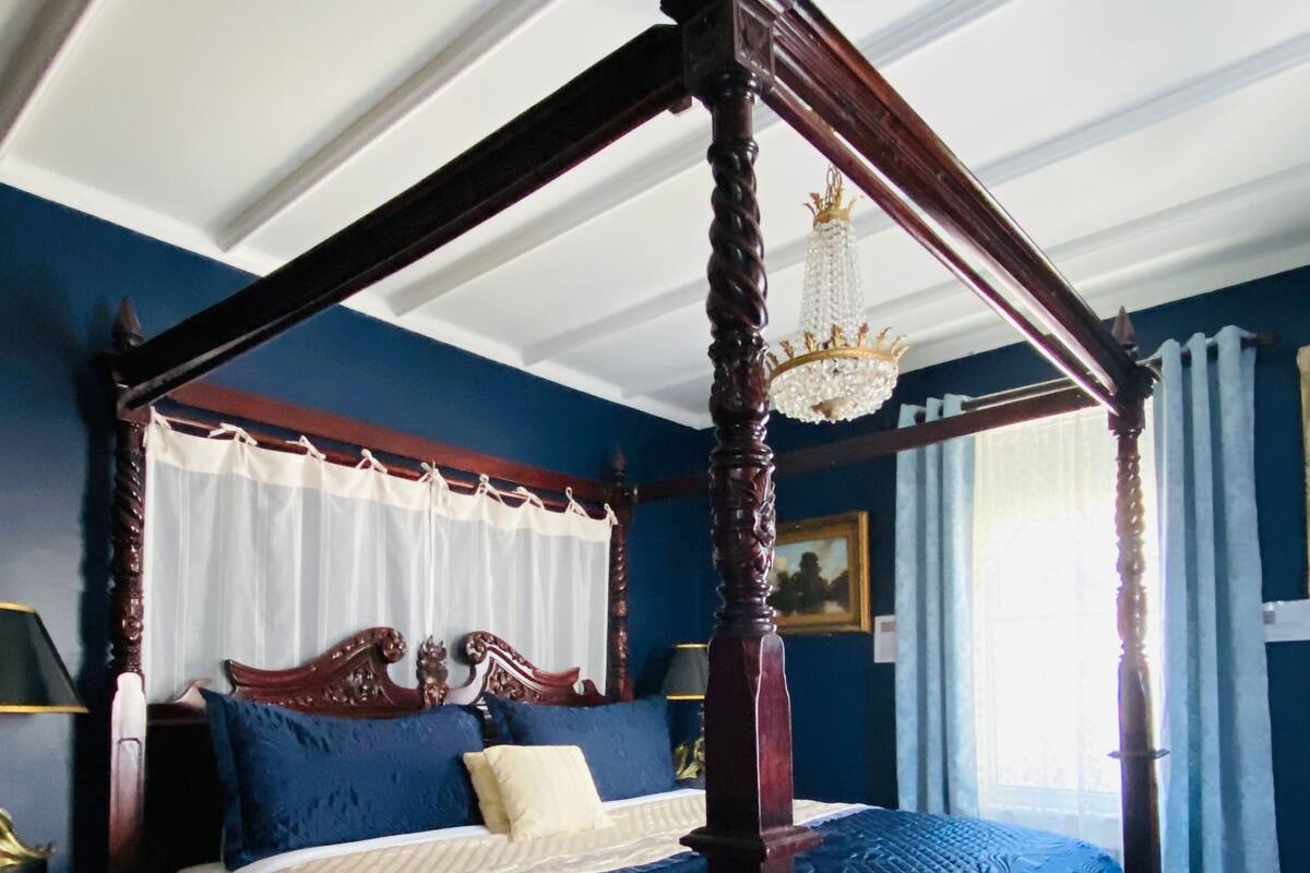 A four-poster bed with a dark wooden frame and a white canopy, a blue upholstered headboard, and a patterned bedspread.